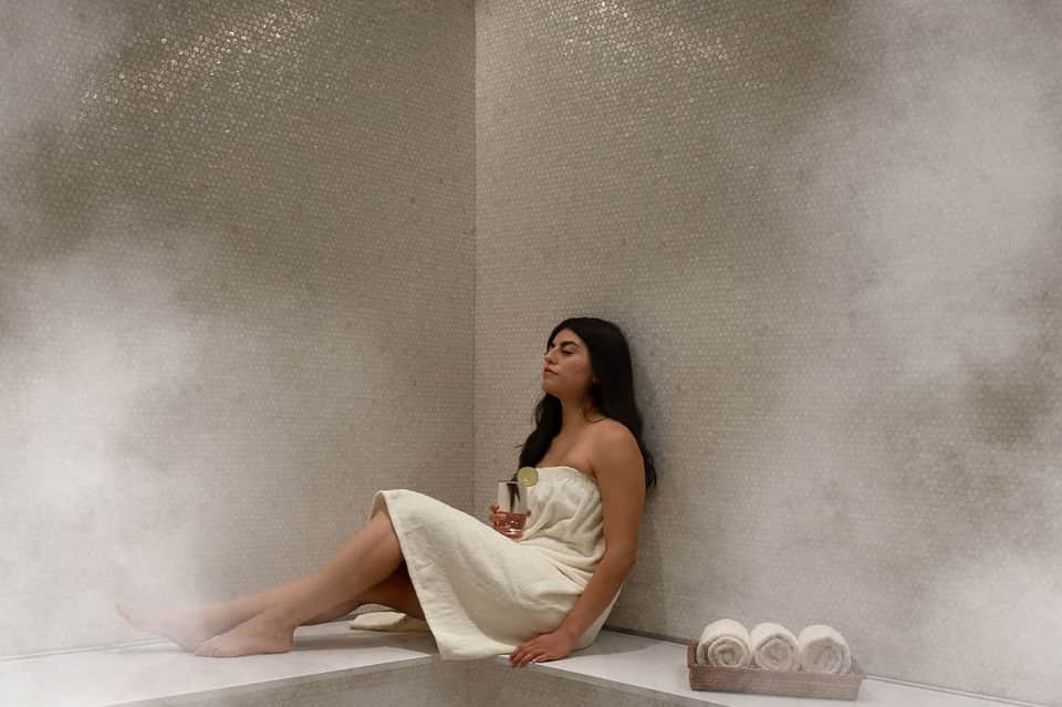 Woman in cream towel wrap sitting in minimalist spa corner with rolled towels and candle