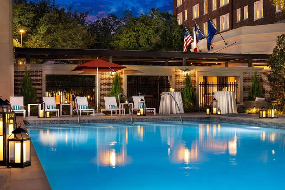 Elegant nighttime pool area with illuminated water, brick buildings, lounge chairs, and umbrellas under blue evening sky