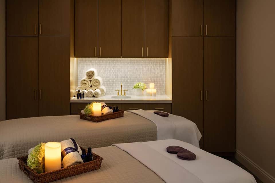 Luxury spa treatment room with two massage tables, lit niche with rolled towels, and dark wood cabinetry