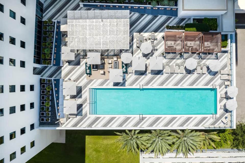 Aerial view of modern rooftop with rectangular pool, lounge chairs, umbrellas, and striped deck surrounded by white buildings