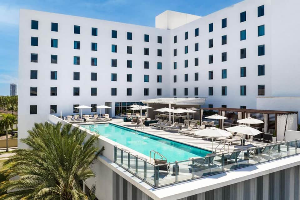 Modern white hotel building with rooftop pool, lounge chairs, and umbrellas overlooking the waterfront