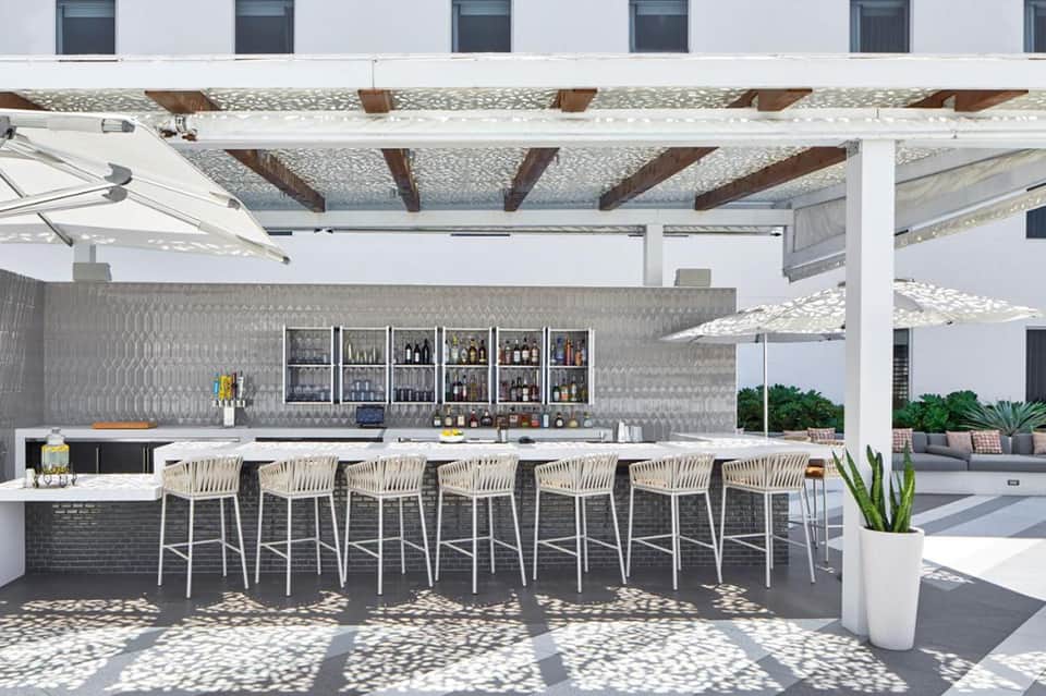 Modern outdoor bar under pergola with white stools, patterned backsplash, and lounge seating in background