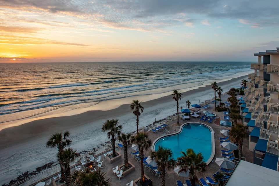Beachfront resort at sunset with multi-story building, pool, palm trees, and sandy beach with ocean waves