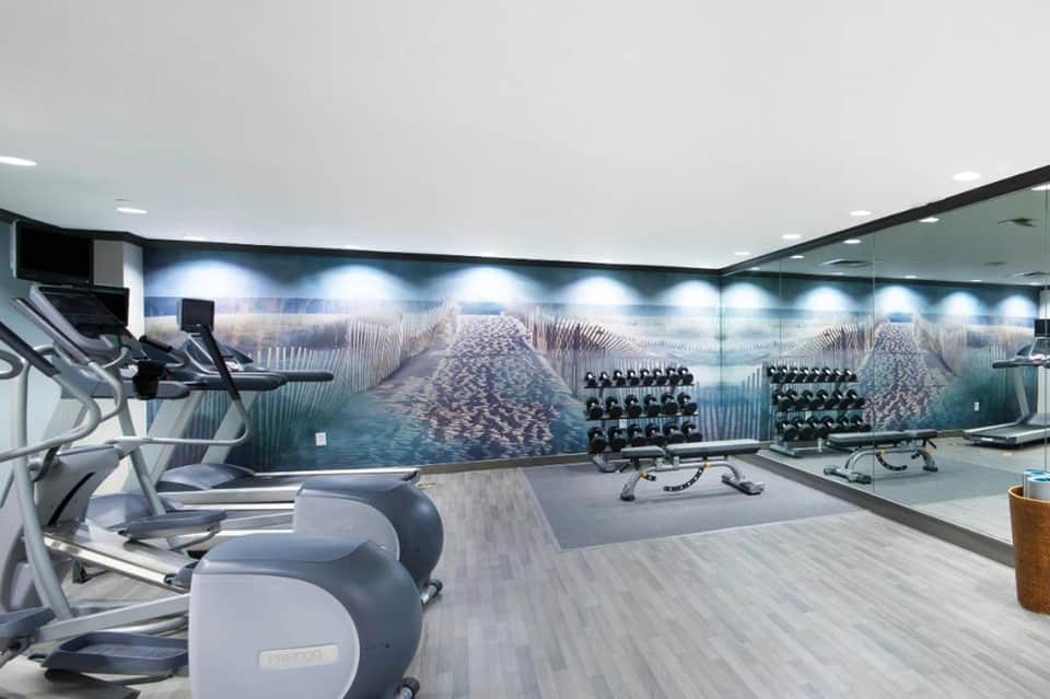 Contemporary fitness center with cardio equipment, dumbbells, and inspirational beach mural on the walls.