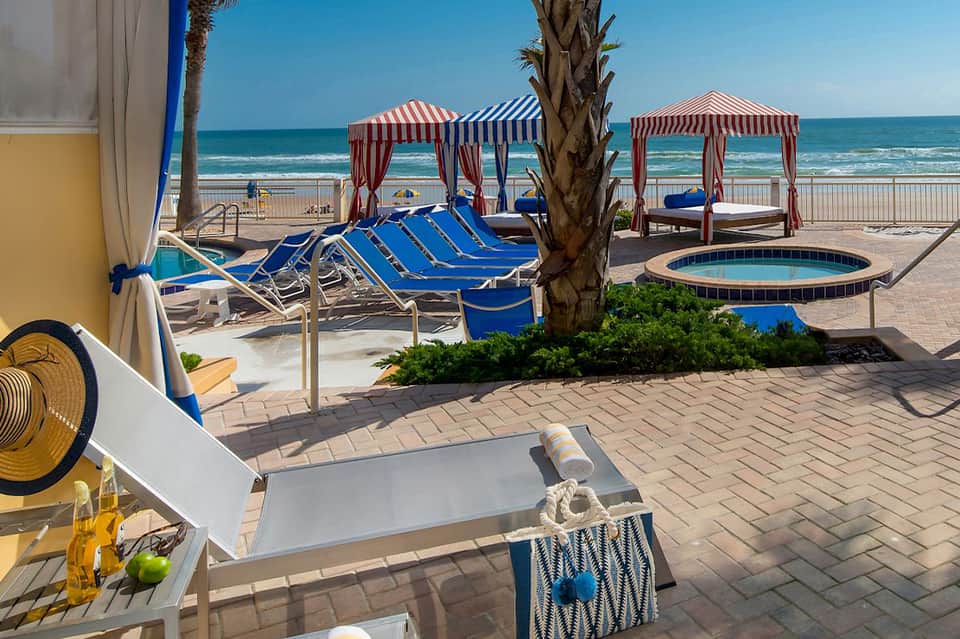 Beach resort patio with blue lounge chairs, striped cabanas, palm tree, and ocean view