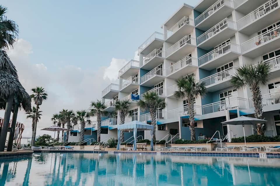 Multi-story beachfront resort with white balconies, palm trees, and swimming pool