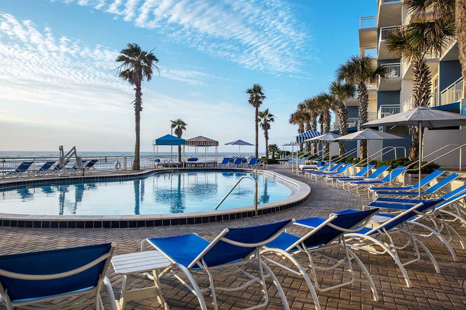 Beachfront resort pool area with blue lounge chairs, palm trees, and ocean view