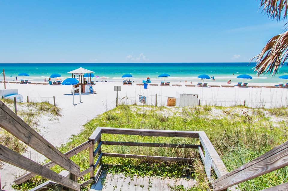 Wooden boardwalk leading to pristine white sand beach with blue umbrellas and turquoise ocean water