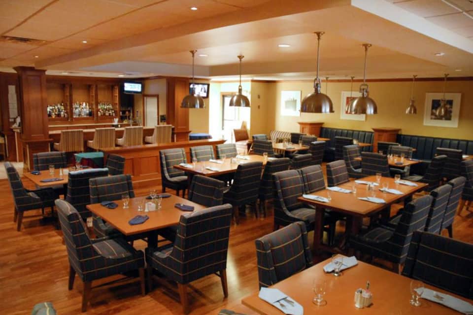 Upscale dining room with wooden bar, multiple tables with plaid chairs, pendant lighting, and warm ambient lighting