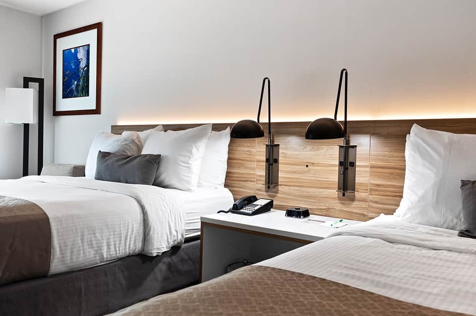 Modern hotel room with two queen beds, wood headboard, and ambient lighting above the headboard