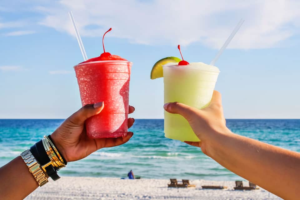 Two colorful frozen cocktails with cherries and straws held against ocean and beach backdrop