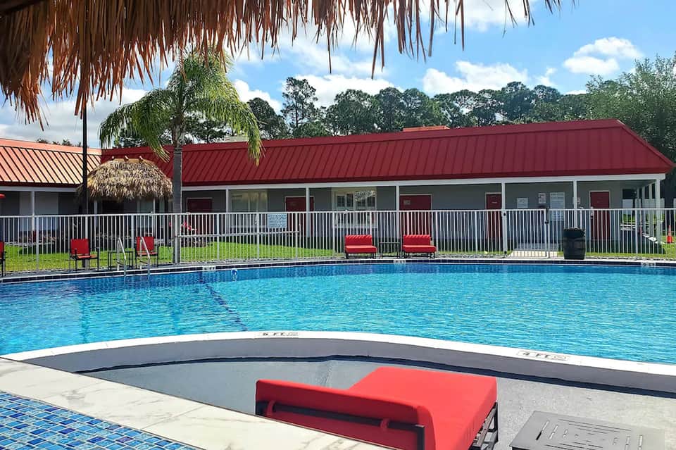 Resort pool with red metal roof building, white railings, lounge chairs, and tropical landscaping under blue sky