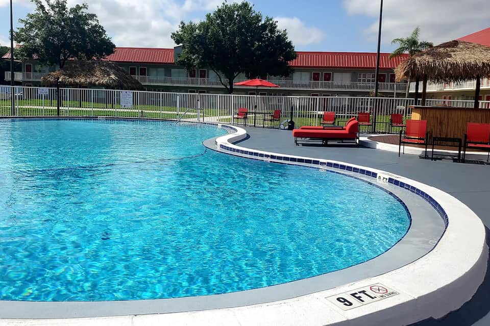 Resort swimming pool with curved white edge, red loungers, palm trees, and residential buildings in background