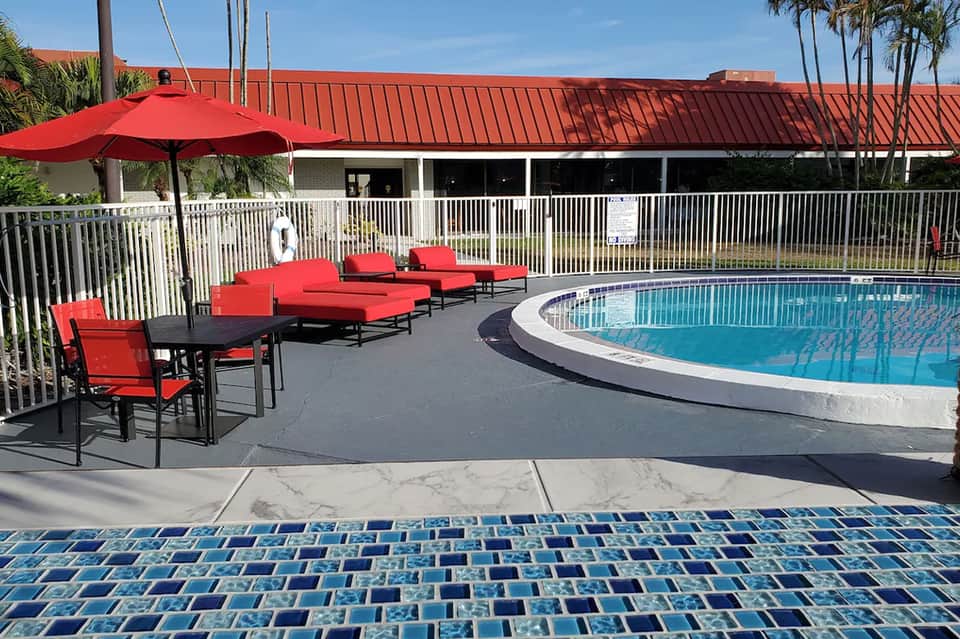 Resort pool area with red lounge chairs, umbrella, and white fencing under clear blue sky