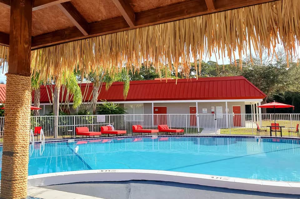 Tropical resort pool with thatched palapa roof, red lounge chairs, and red-roofed building with white railings