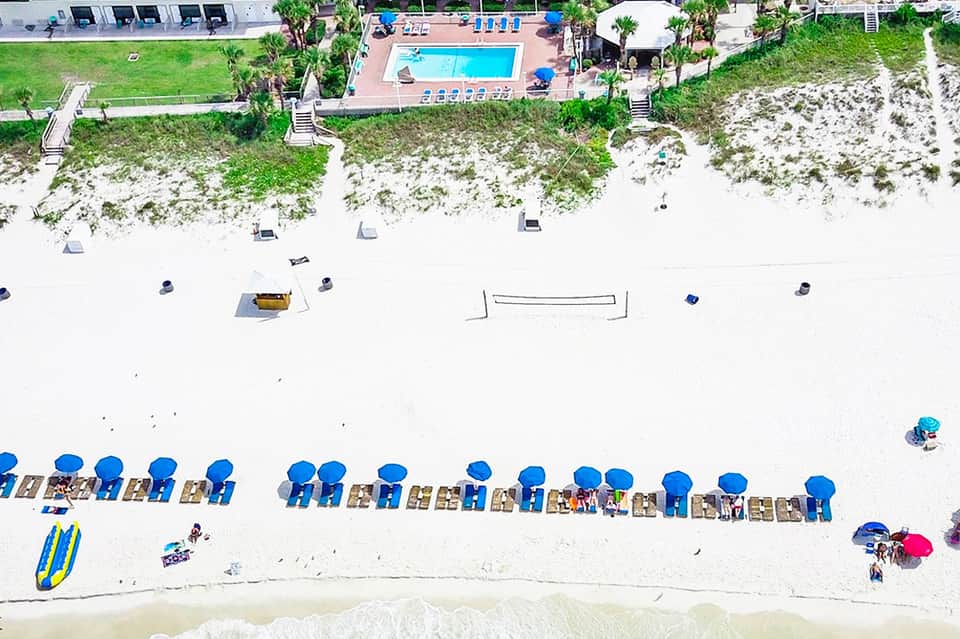 Aerial view of beachfront resort with blue umbrellas, loungers, pool, and sandy beach with ocean