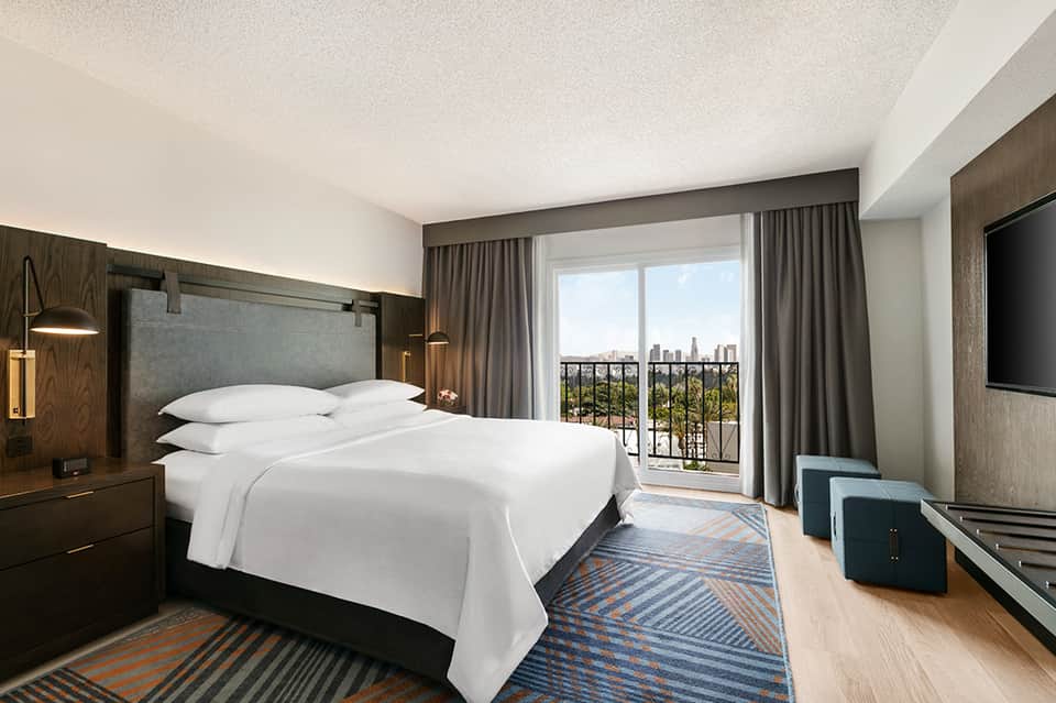 Modern hotel bedroom with dark wood furnishings, white bedding, and floor-to-ceiling windows overlooking a city skyline