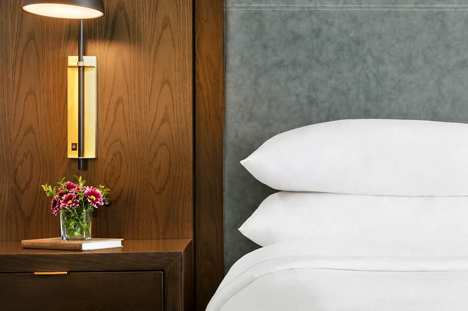 Luxury hotel bedside table with elegant lamp, fresh flowers, and white pillows on bed with gray headboard