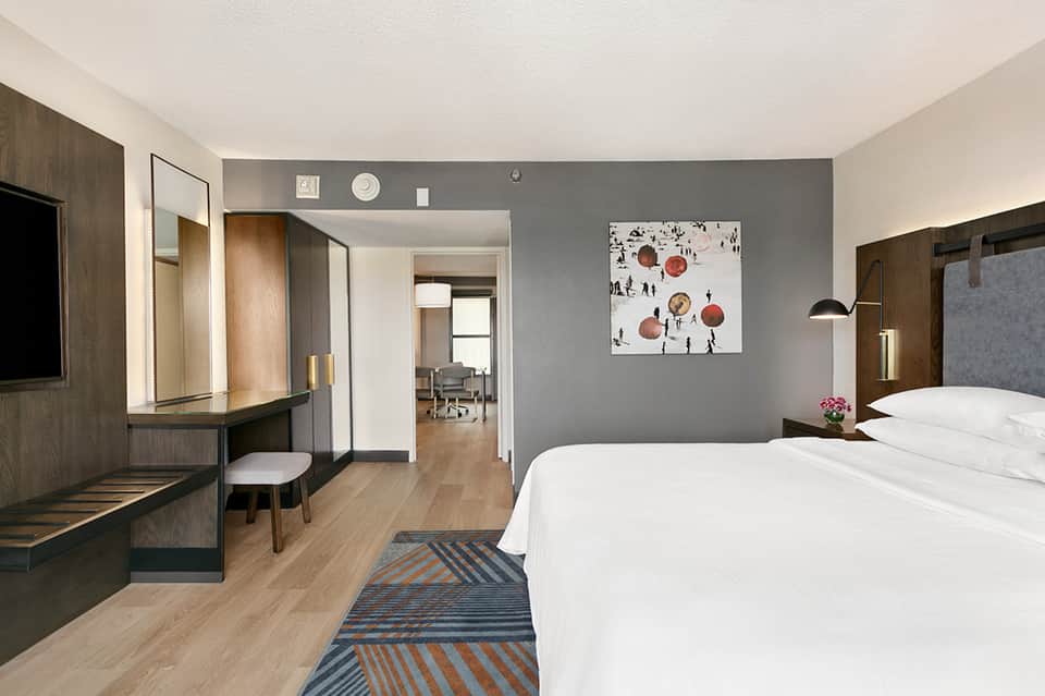 Modern hotel bedroom with gray accent wall, wooden vanity, white bedding, and access to work area