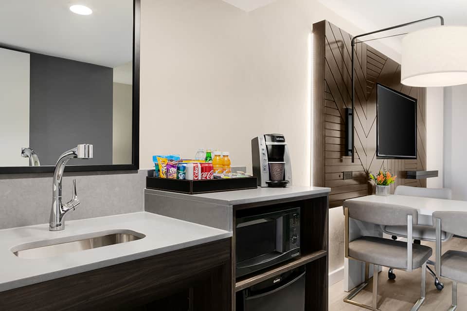Modern hotel room kitchenette with white countertop, dark cabinetry, sink, microwave, and beverage station with snacks
