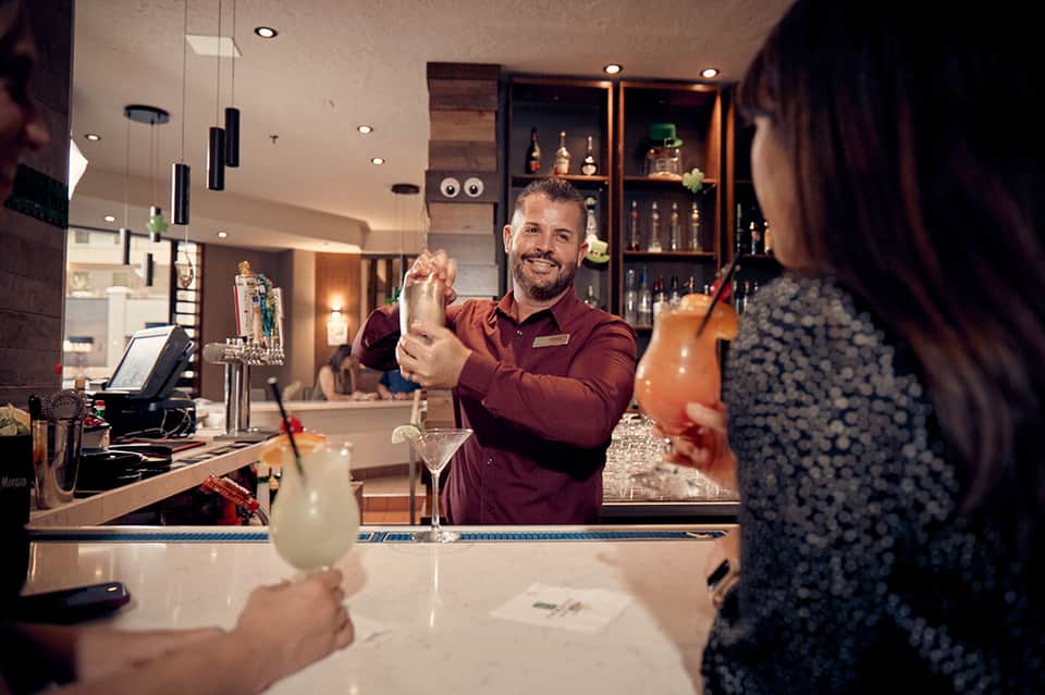 Bartender in maroon shirt preparing cocktails at modern hotel bar with shelving and warm lighting