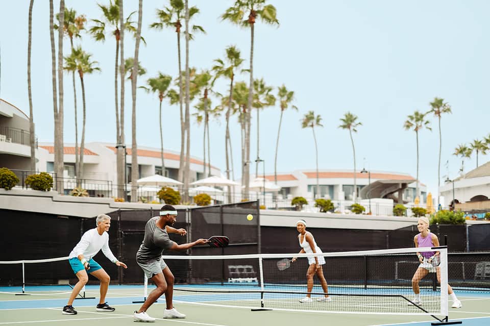 Group playing pickleball on outdoor court with palm trees and white hotel buildings in background