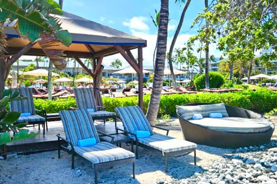 Beachfront lounge area with striped blue cushioned chairs, wooden pergola, and palm trees overlooking ocean