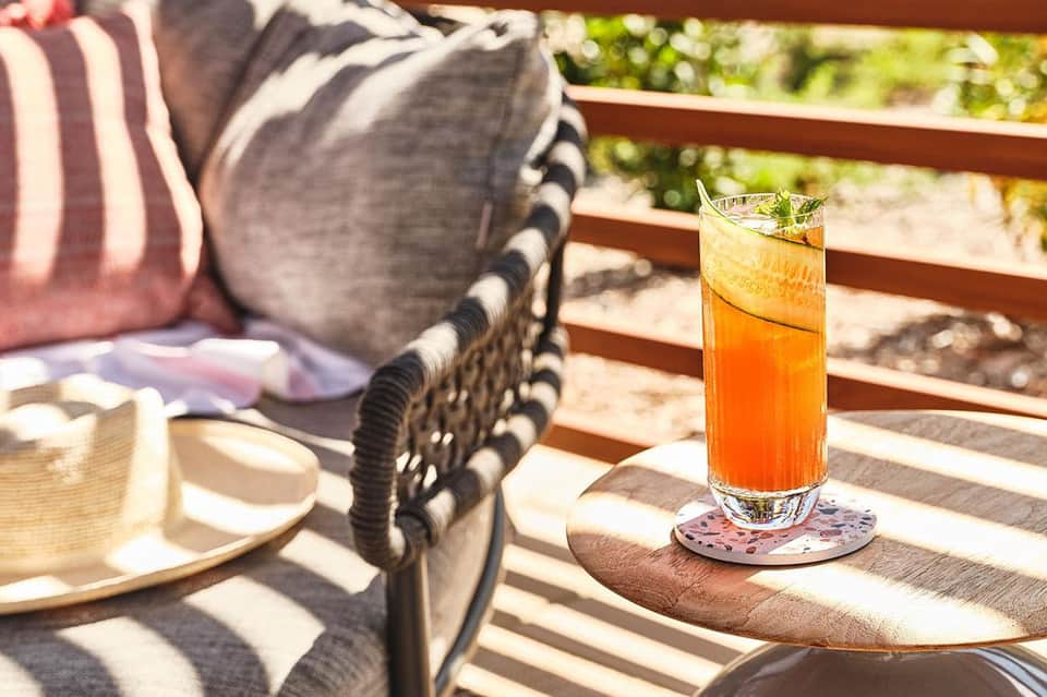 Iced beverage on wooden table beside lounge chairs on sunny patio with striped cushions