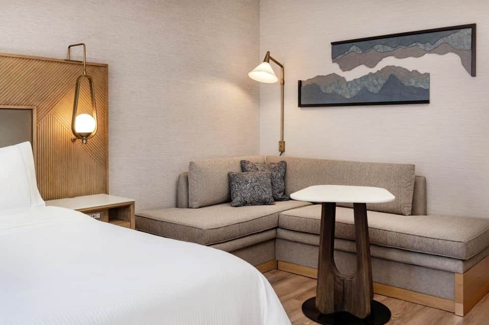 Contemporary hotel room with white bed, gray upholstered sofa, wooden side table, and mountain artwork