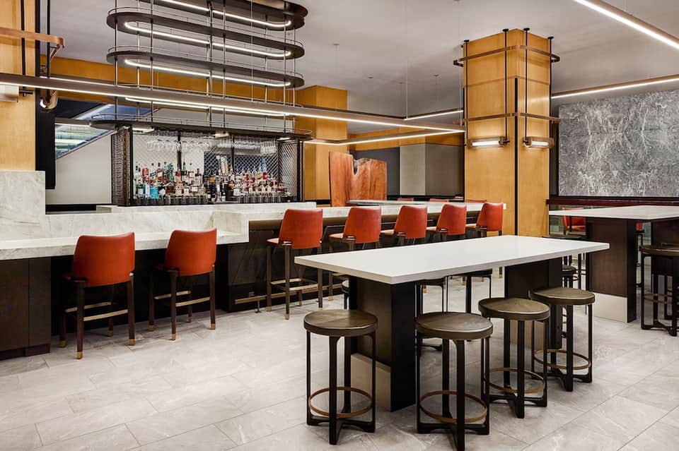 Modern hotel bar and lounge with orange accent chairs, wooden beams, and marble countertops