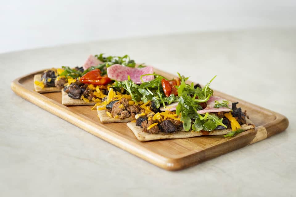 Four open-faced sandwiches on wooden board with ground meat, yellow cheese, greens, and pickled vegetables