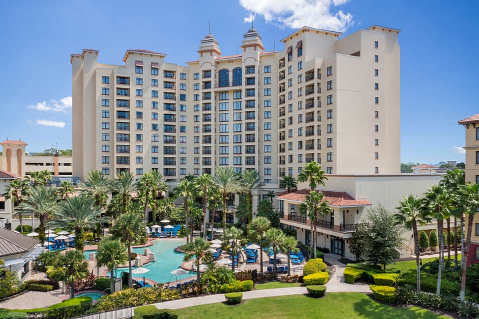 Large beige resort building with palm trees and curved swimming pool surrounded by landscaped grounds
