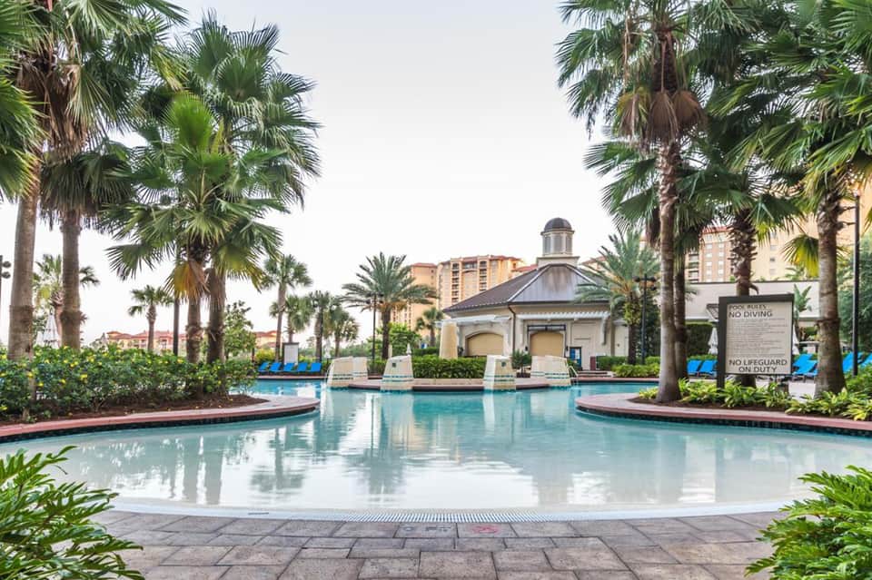 Luxurious resort pool surrounded by palm trees with colonial-style pavilion and high-rise buildings in background