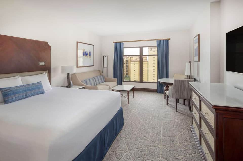 Spacious hotel room with king bed, seating area, and balcony overlooking city buildings