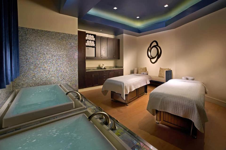 Spa treatment room with two massage beds, hot tubs, and wooden cabinetry with ambient lighting