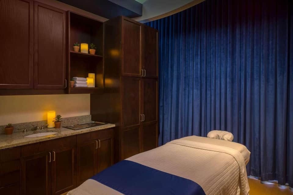 Spa treatment room with dark wood cabinetry, massage bed with cream linens, and blue curtains