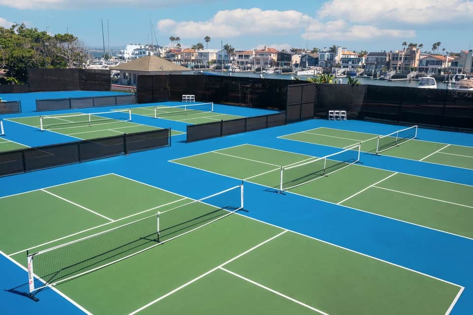 Waterfront tennis courts with blue and green playing surfaces overlooking a coastal neighborhood with boats and palm trees