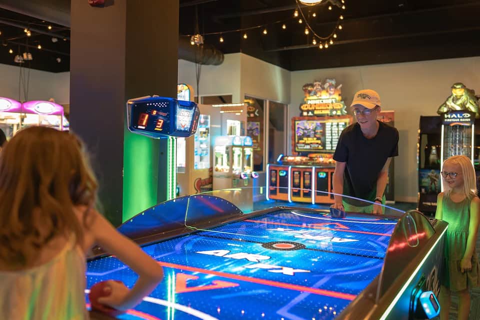 Interactive arcade game area with glowing blue air hockey table and vintage game machines in background