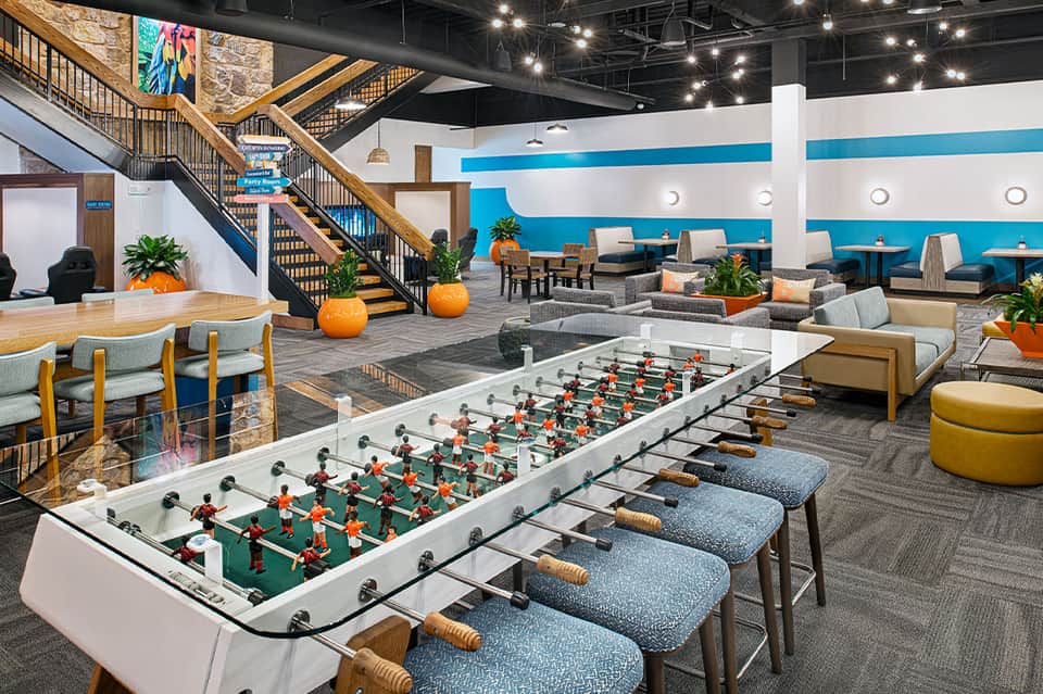 Modern hotel recreation area with foosball table, seating areas, and wooden staircase with blue accents