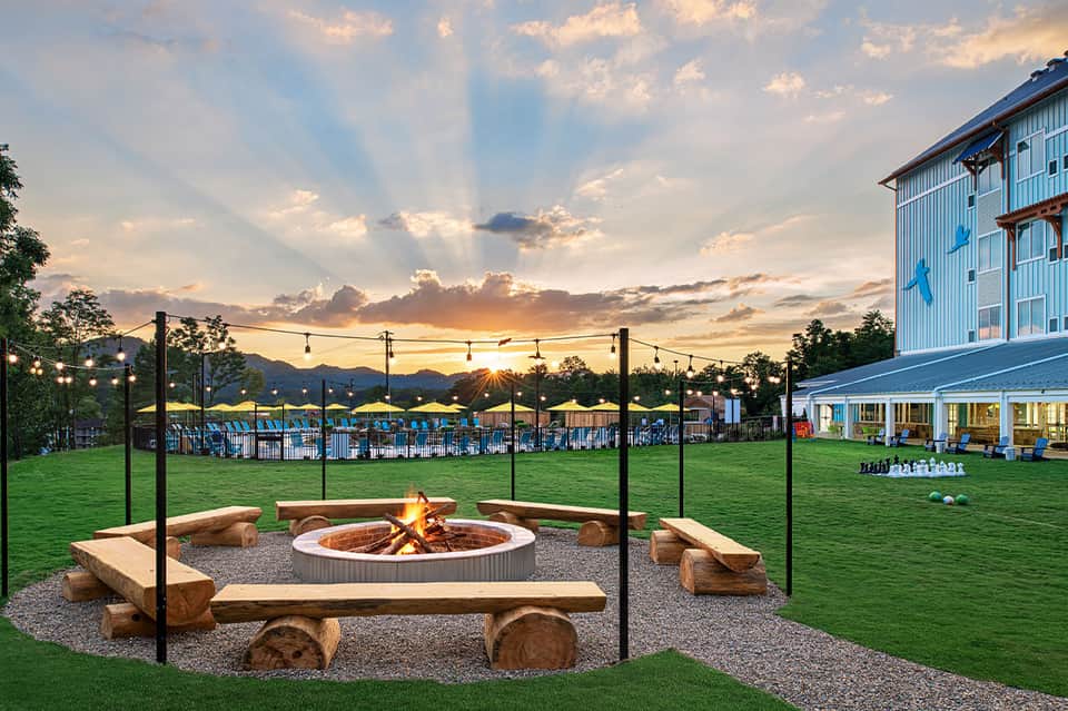 Outdoor fire pit with wooden bench seating surrounded by manicured lawn, mountains, and blue building at sunset