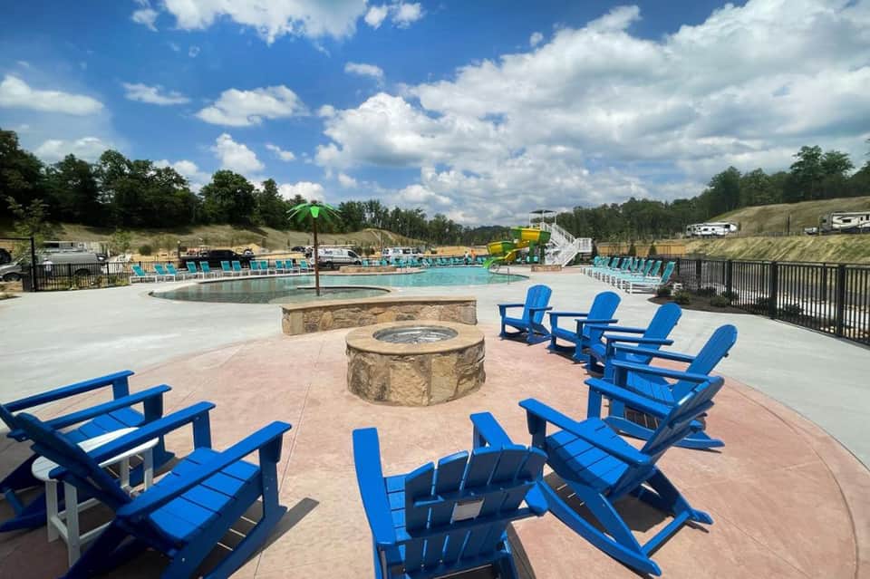 Resort fire pit area with blue Adirondack chairs surrounding stone fireplace, pools and green hills visible in background