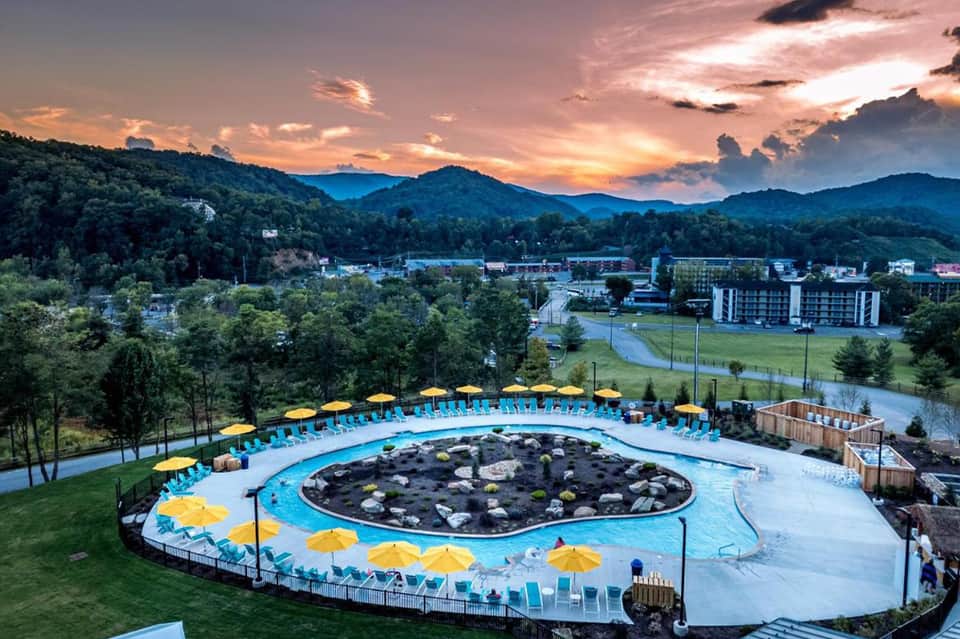 Circular hot spring pool with rocky center surrounded by yellow umbrellas at sunset, mountain backdrop