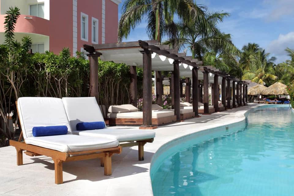 Luxury resort pool area with wooden lounge chairs, pergola structures, and tropical palm trees