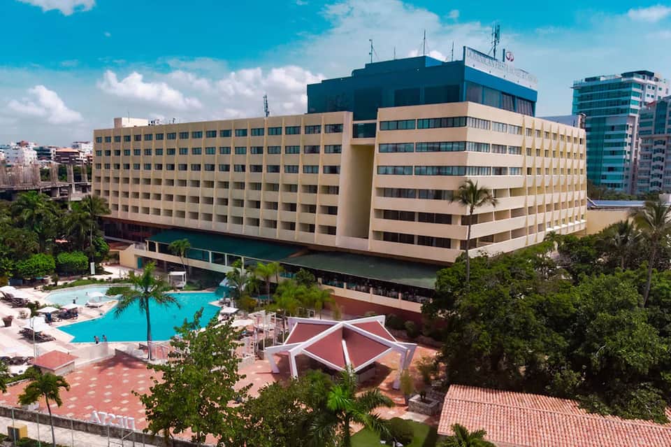 Beige multi-story hotel with curved architecture and bright blue swimming pool surrounded by palm trees