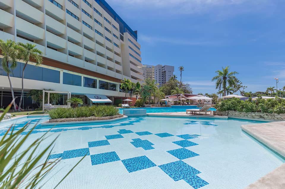 Resort pool with blue and white checkered tile pattern, palm trees, and multi-story hotel building under clear sky