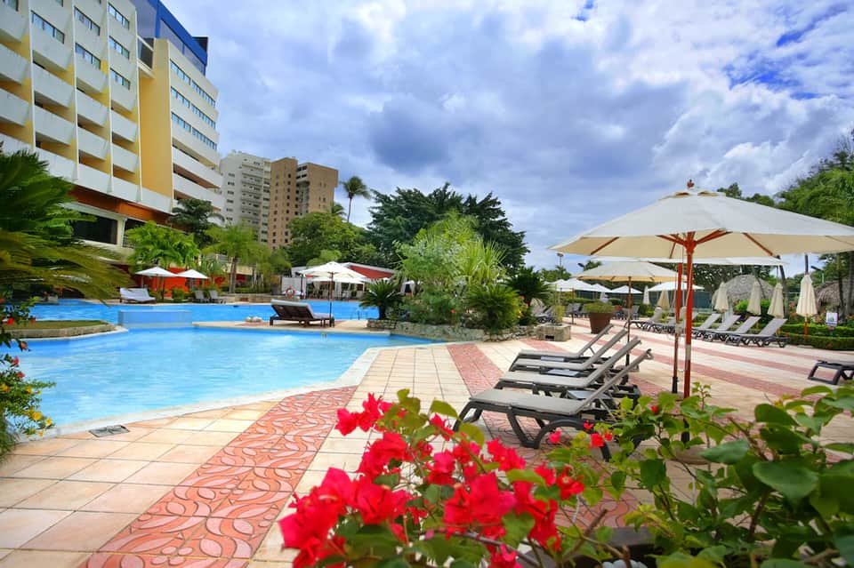 Resort pool area with blue water, lounge chairs, red umbrellas, tropical plants, and yellow apartment building in background