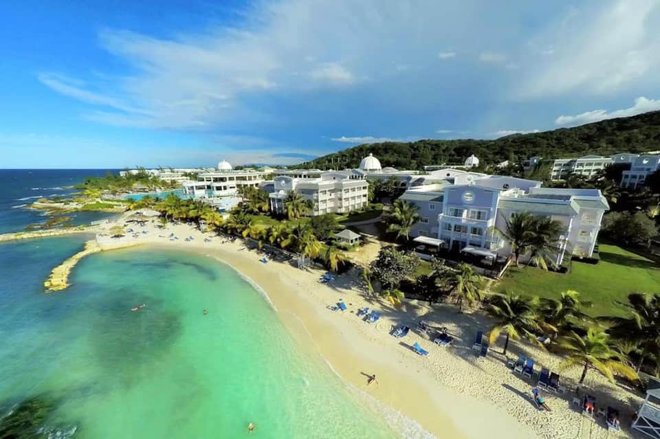 Aerial view of beachfront resort with turquoise waters, white buildings, palm trees, and sandy beach with lounge chairs