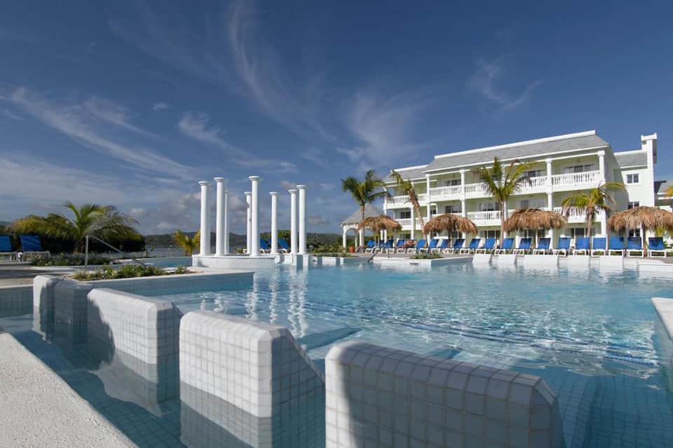 Luxurious waterfront resort with white columns, pool, tiki-roofed cabanas, and palm trees under blue sky