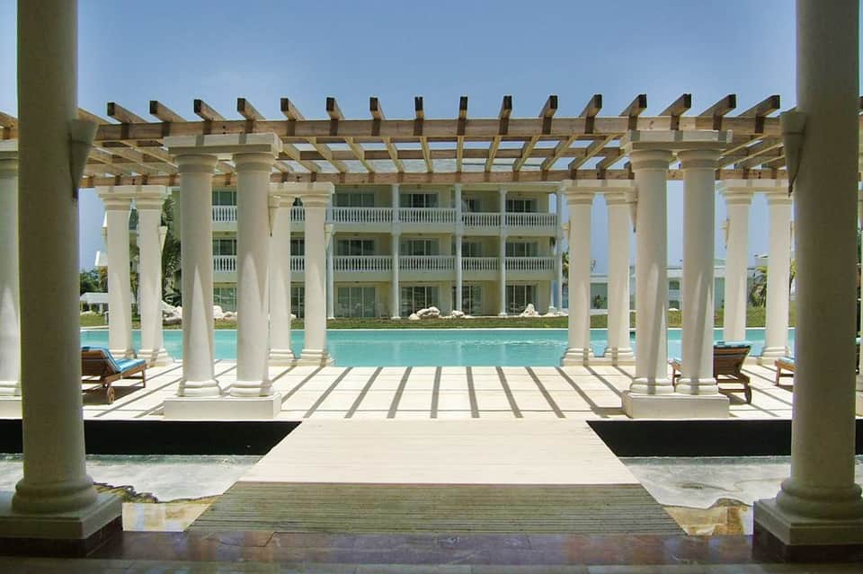 Modern white pergola structure framing turquoise pool with neoclassical resort building backdrop