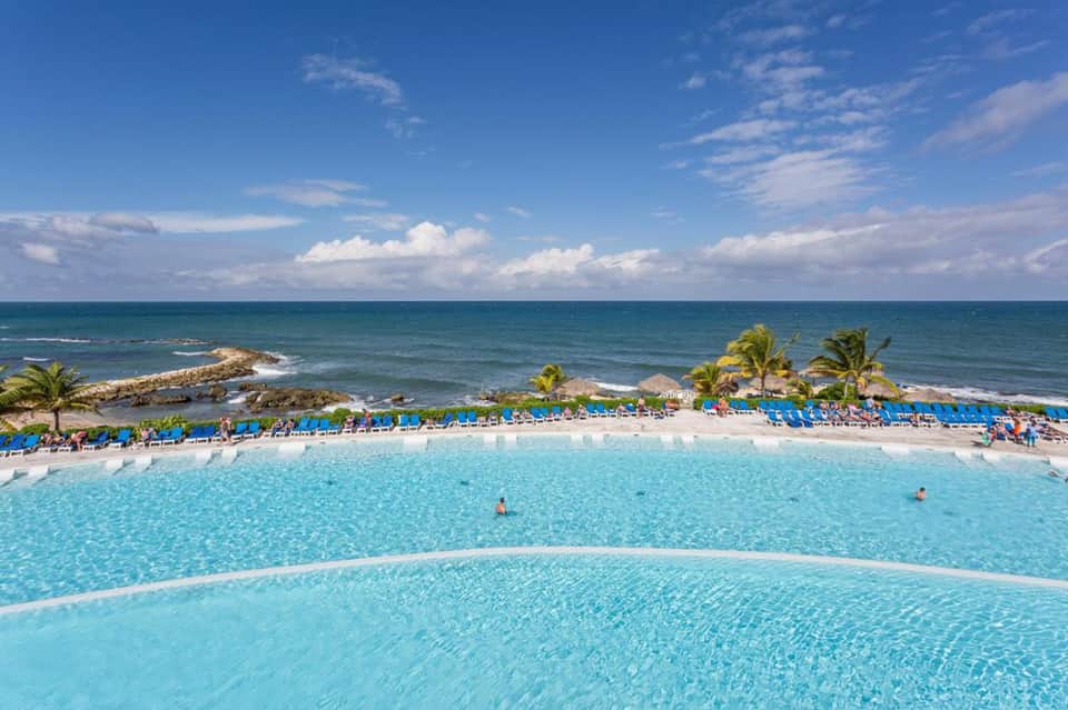 Resort pool with turquoise water, lounge chairs, palm trees, and ocean view in background